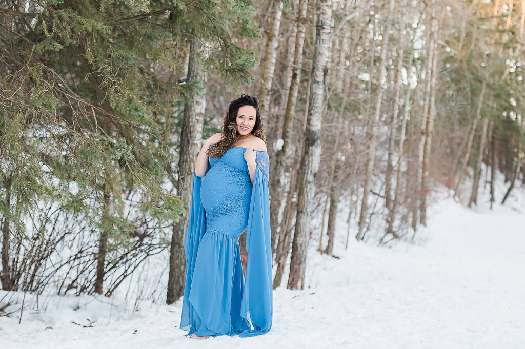 Photograph of an Edmonton mother wearing a blue floor length flowy maternity dress with long sleeves in a winter setting at Mill Creek Ravine at an Edmonton Maternity Photography session