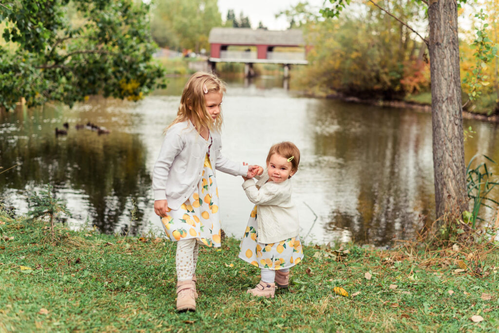 Two sisters playing at Woodbridge Farm in Sherwood Park during a family photo session with the pond in the background