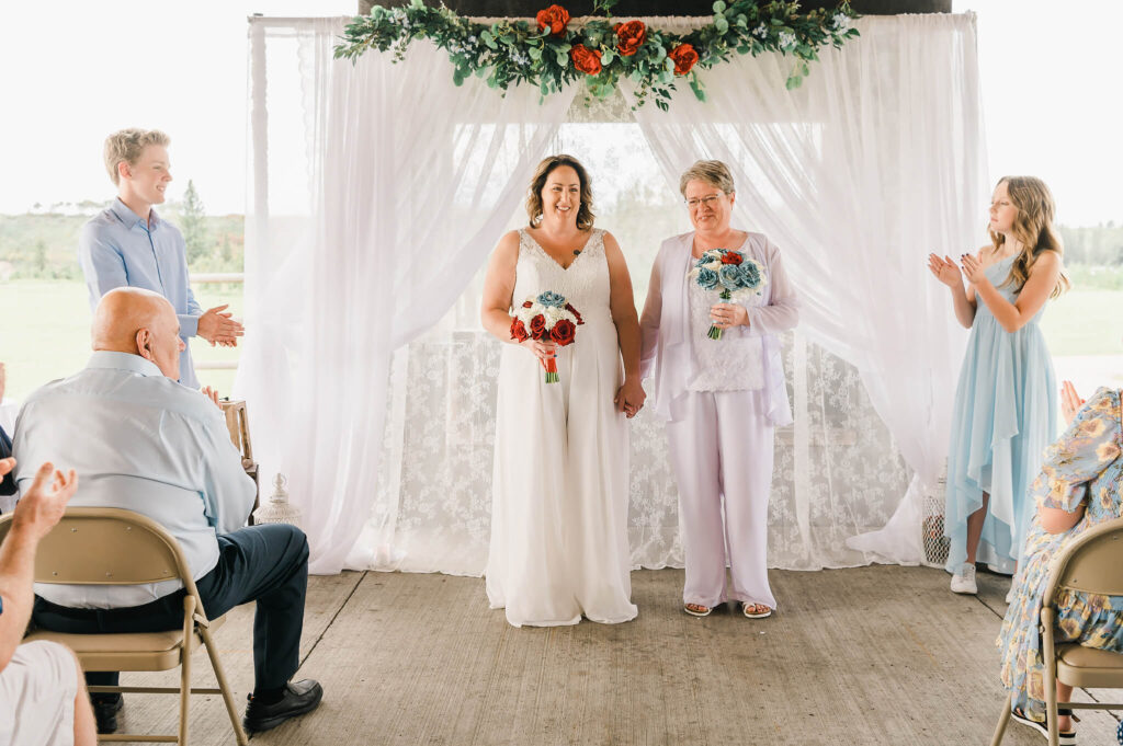 Two brides during a procession at West River's Edge with the fireplace covered by sheer lace and flowers 