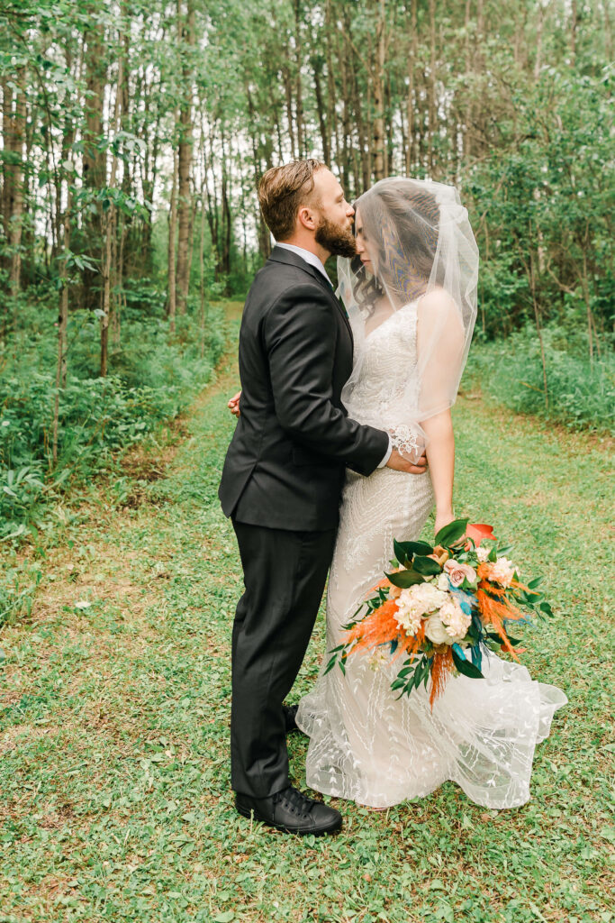 A groom kisses his bride with her veil over her head in a fine art editorial pose while her bouquet is down by her side