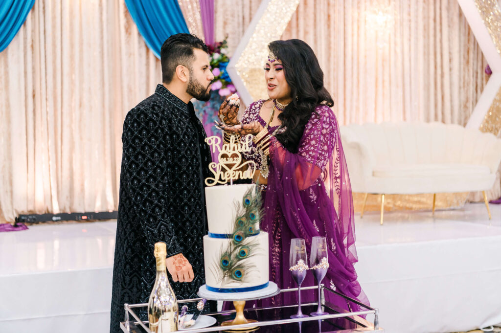 An east indian bride wearing a purple lehenga and a groom wearing a black East Indian attire feeding each other cake during the cake cutting at Royal Palace, Edmonton