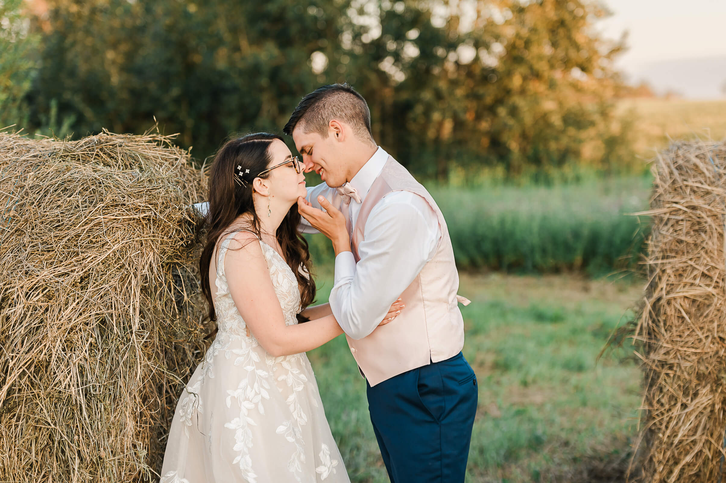 An Albertan wedding couple exchanges a soft moment: groom in pink vest and white shirt holding bride's jaw before giving her a kiss by the haybales