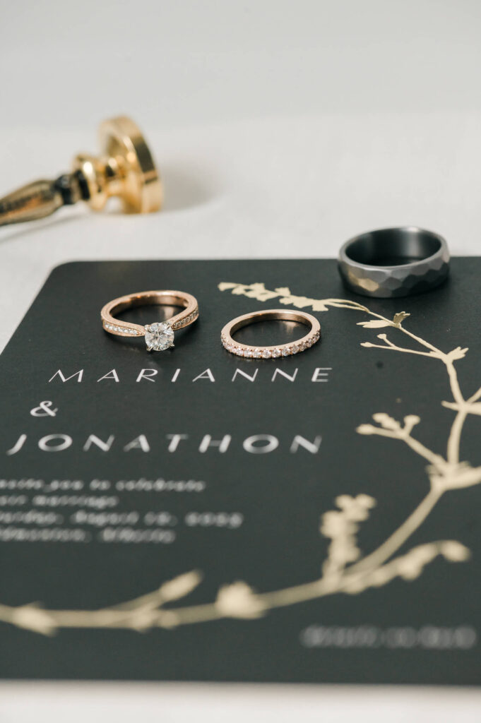 Flat lay details of wedding rings & engagement ring and wedding invite done by Cynthia Priest Photography. Wedding invite is black with gold tree branch with white letterings. 