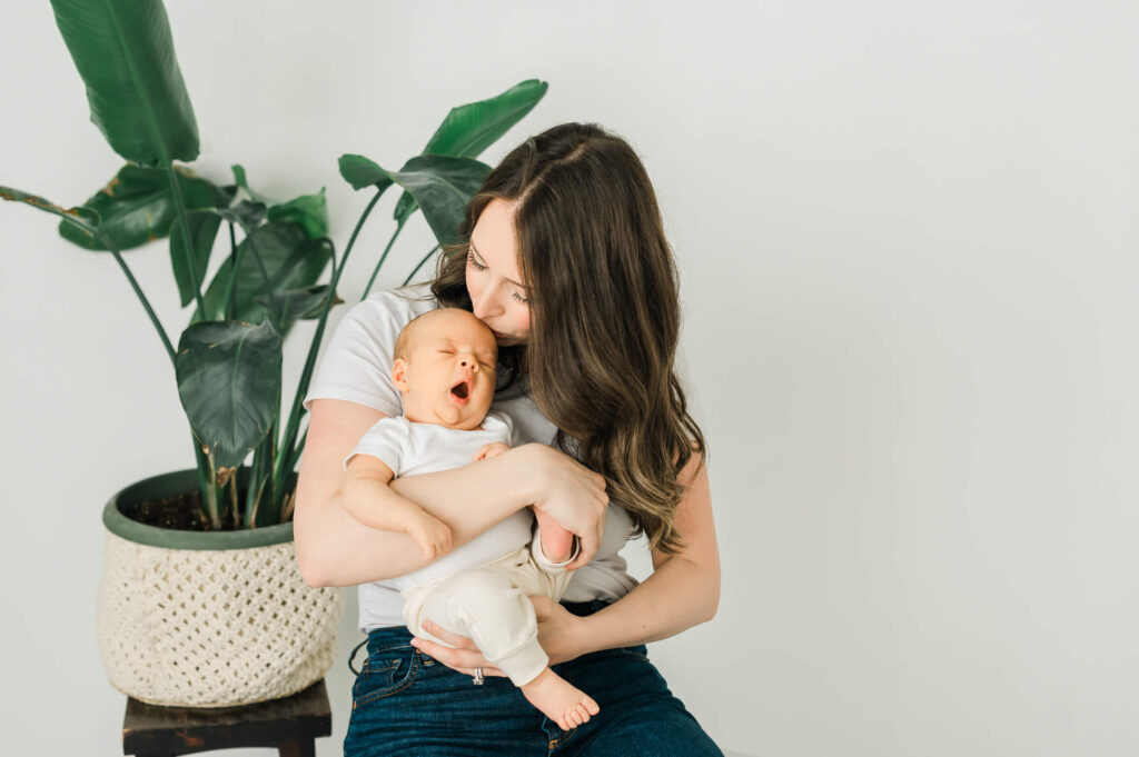 An Edmonton mama in solid white shirt and blue denim pants holding her newborn at Cynthia Priest Photography's studio with a monsterra plant behind her