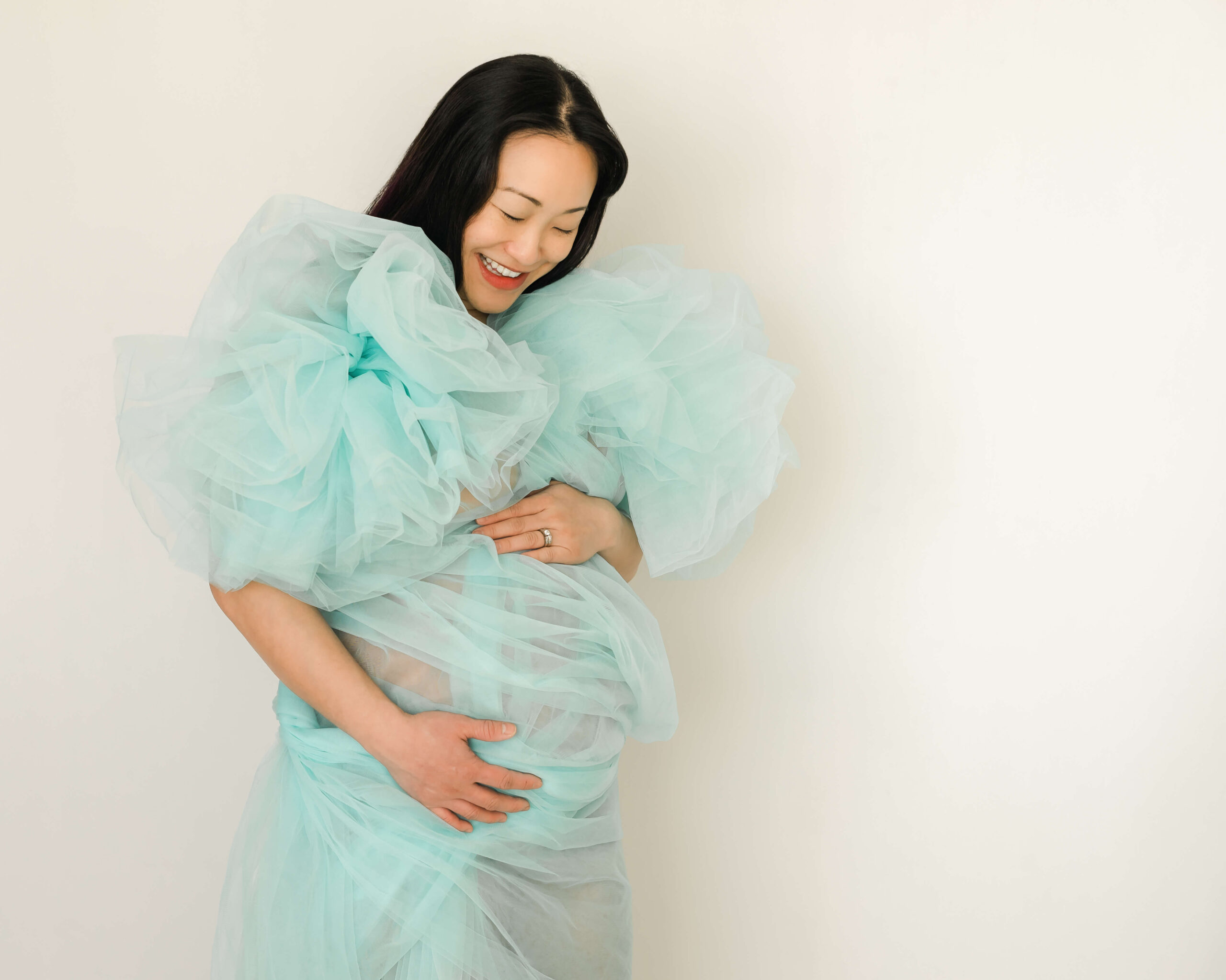 An Asian Edmonton Mom smiling in a couture blue dress holding her baby bump in anticipation and joy in an article talking about Oliver Chiropractic Wellness Clinicc