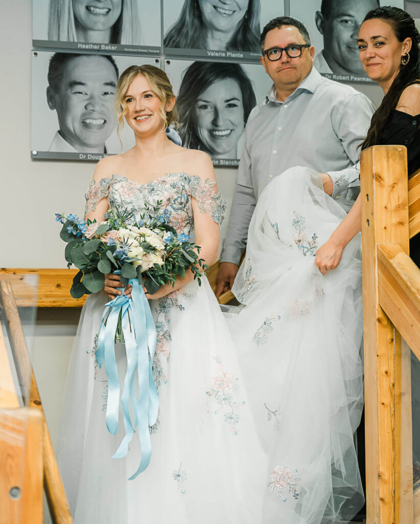 An Edmonton wedding planner Sevy Fays holding a dress for a bride in a floral bridal dress holding a bouquet while walking with her father down the stairs before the ceremony