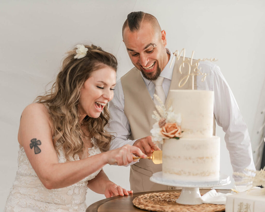 Bride and groom laughing together while cutting their two-tier wedding cake