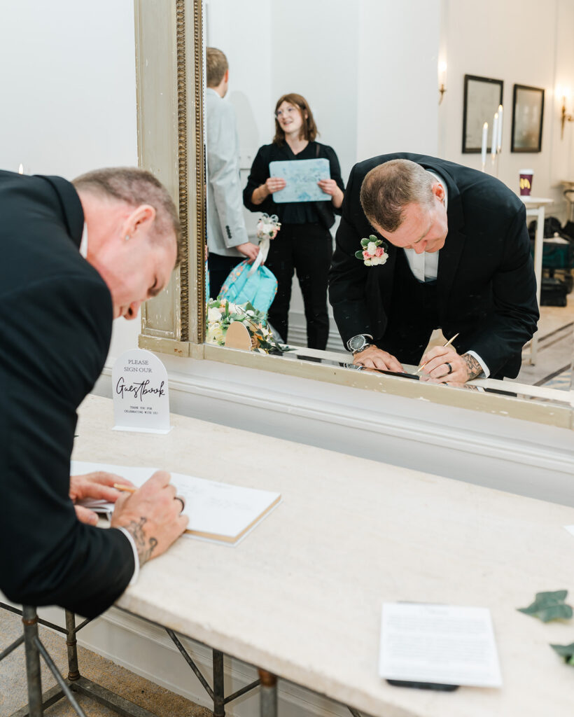A wedding guest signing a guestbook while Meg Wolodko from Hickory and Rose talks to another wedding guest in the background
